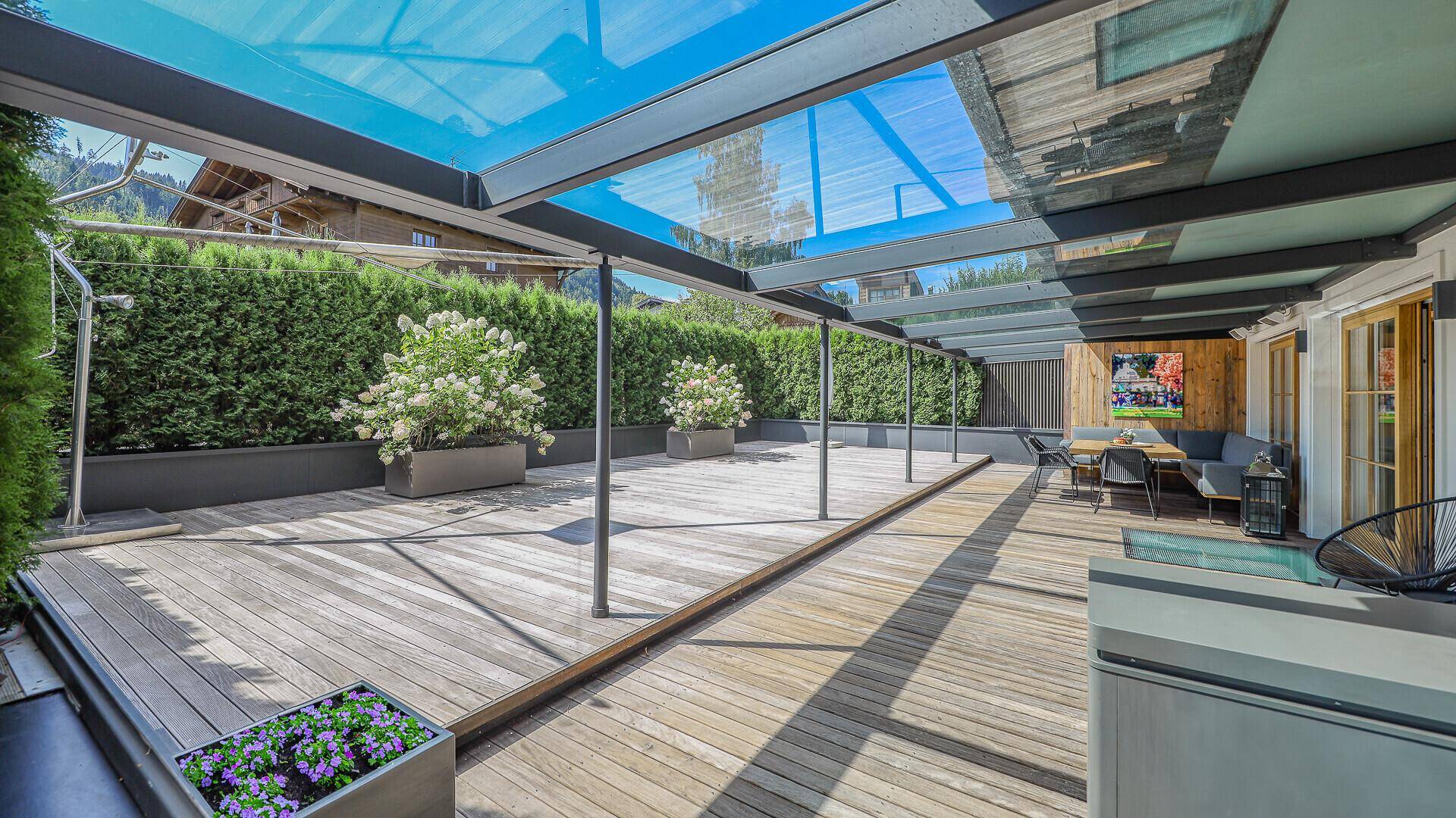 Überdachte Holzterrasse mit Glasdach, umgeben von Hecken und Pflanzen, ideal zum Entspannen.