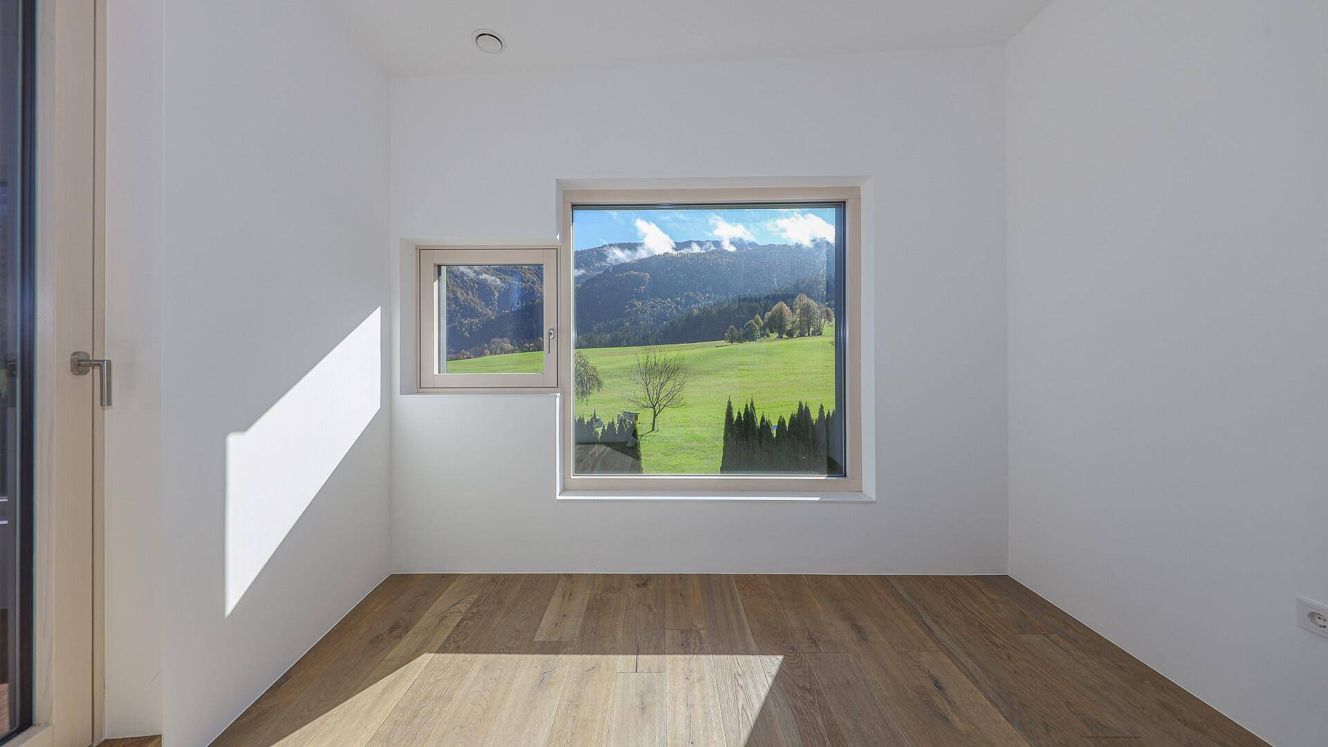 Leeres Zimmer mit Holzboden und Fenstern, die einen weiten Blick auf die Natur ermöglichen.