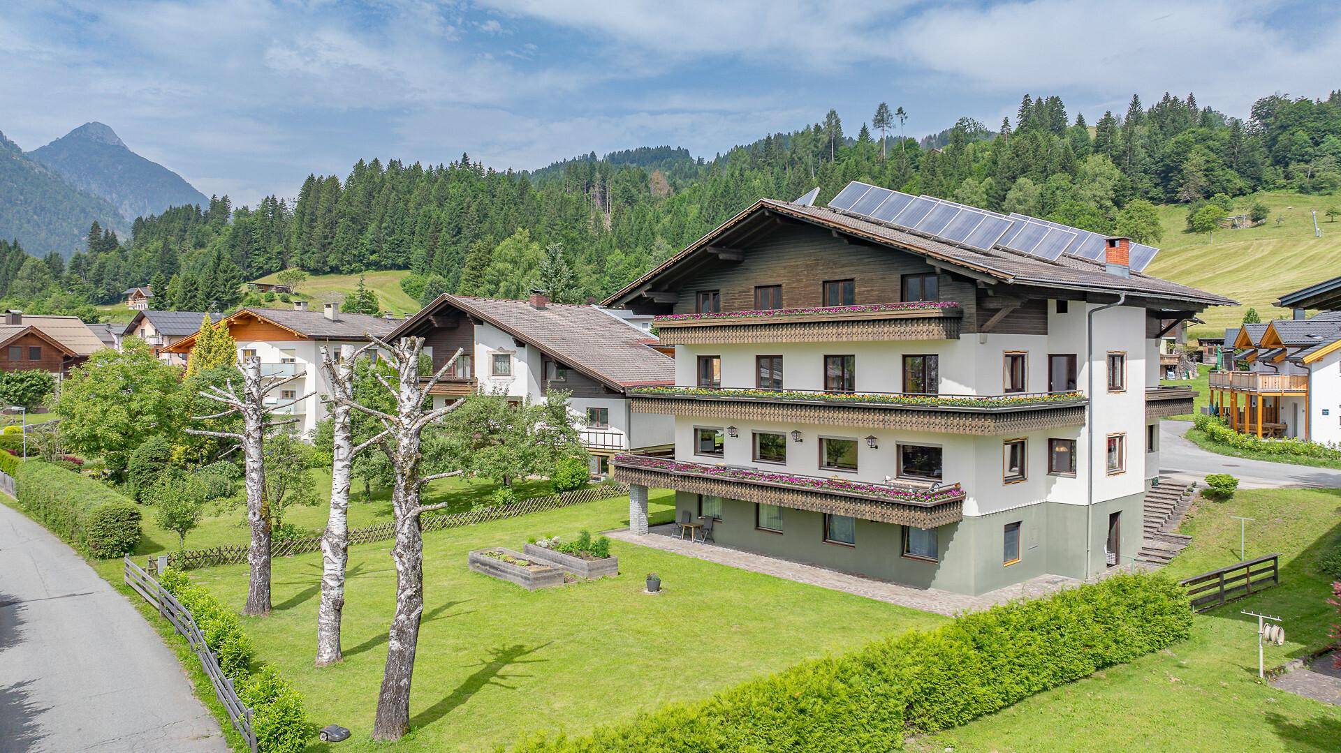 Mehrstöckiges Gebäude mit weißer Fassade, grünen Akzenten und Solarpaneelen in einer Berglandschaft.