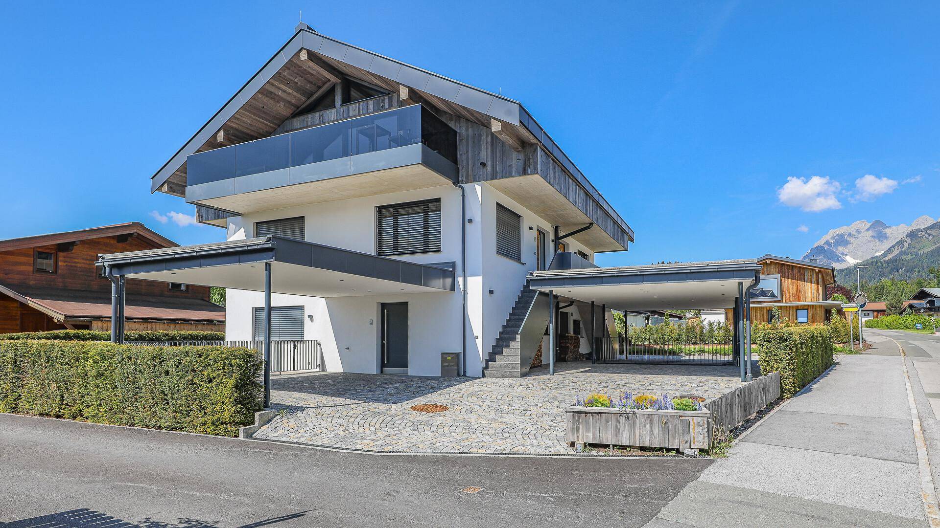 Modernes Haus mit Carport, Balkon und Bergblick unter blauem Himmel an einem sonnigen Tag.