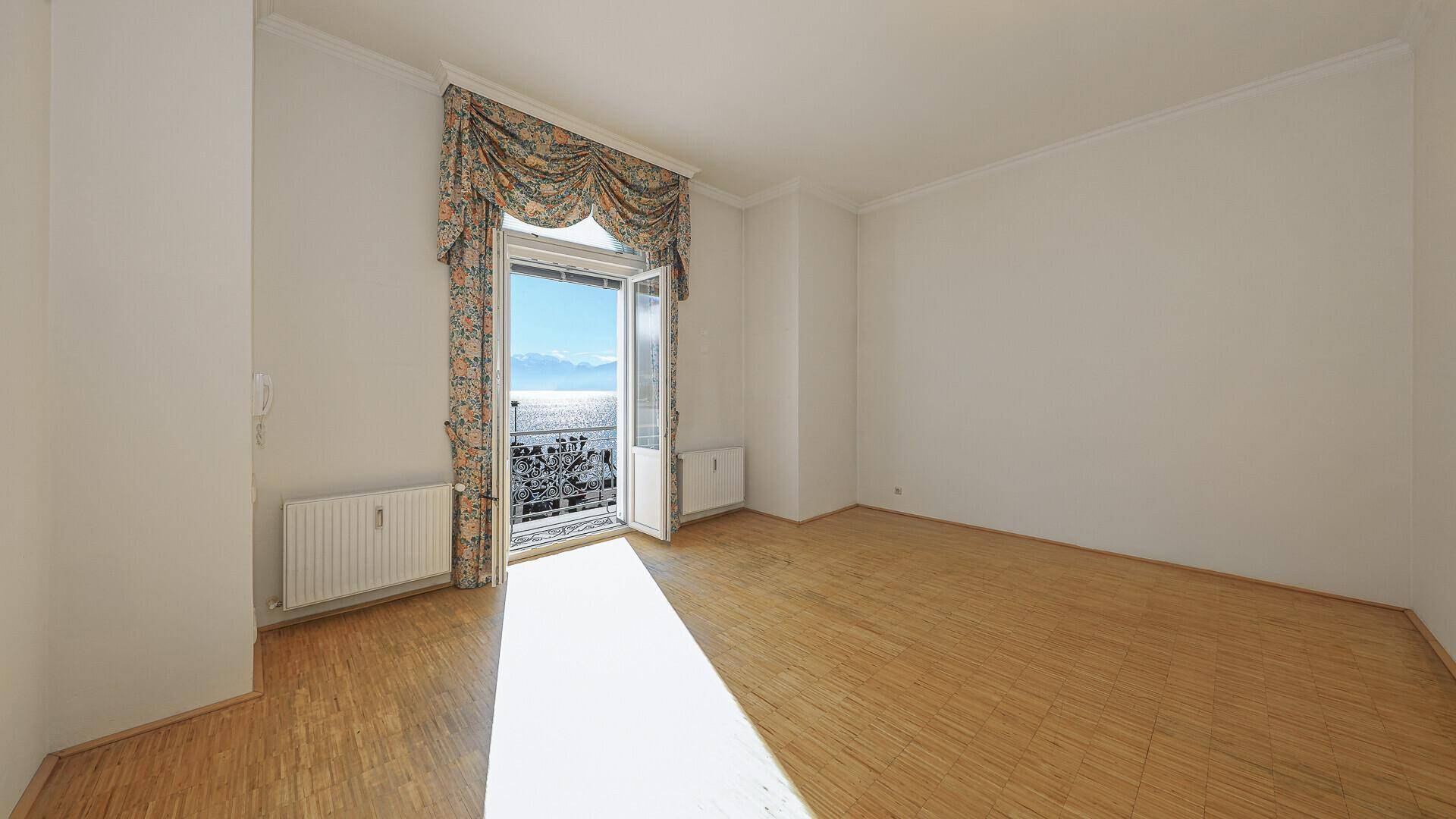 Sonniger, leerer Raum mit Parkettboden und französischem Balkon mit Seeblick.