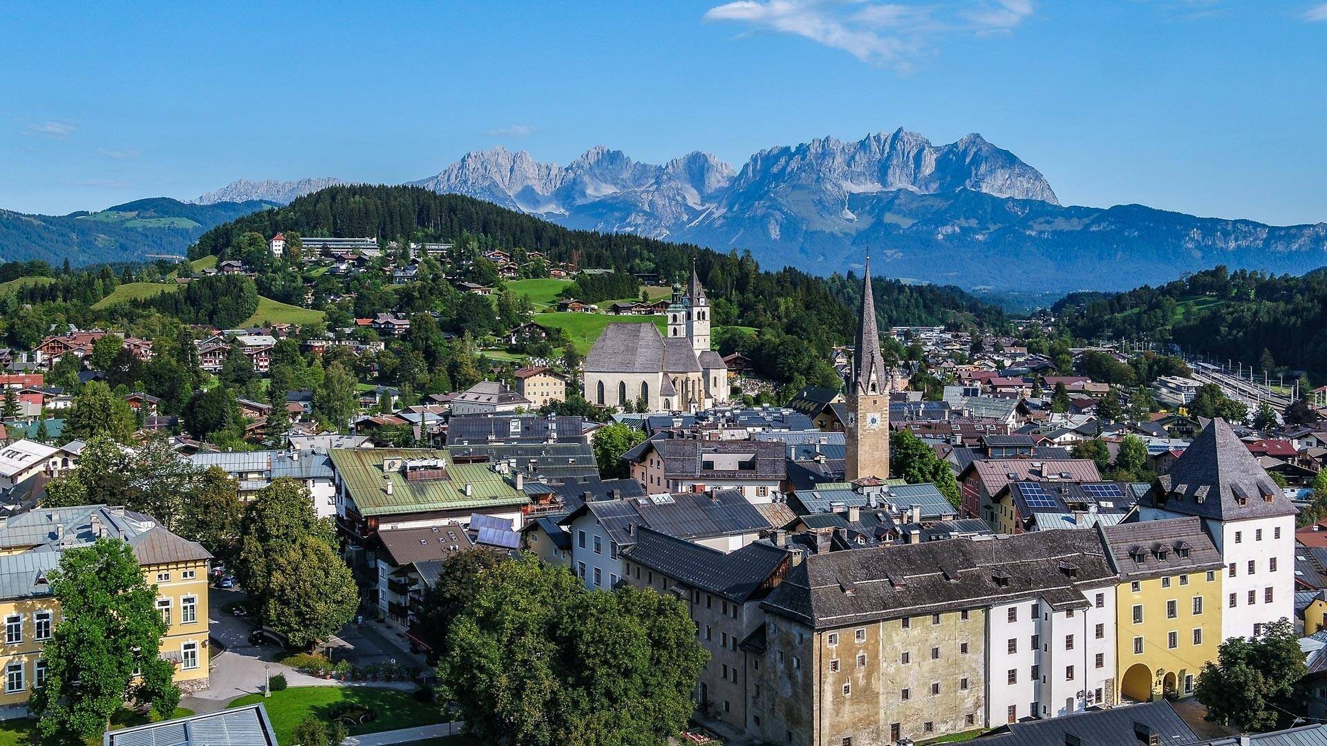Panoramablick auf eine malerische Stadt mit Kirchtürmen und grünen Hügeln vor einer beeindruckenden Bergkulisse.