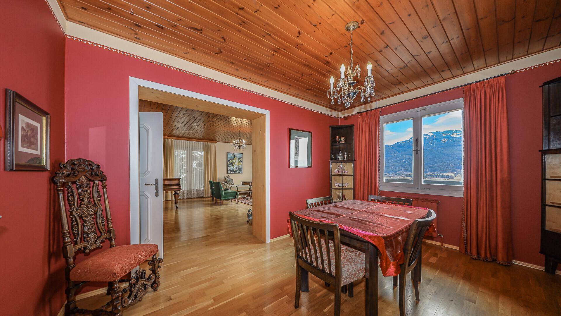 Traditionelles Esszimmer mit roter Wandfarbe, Holzdecke und Bergblick durch das Fenster.