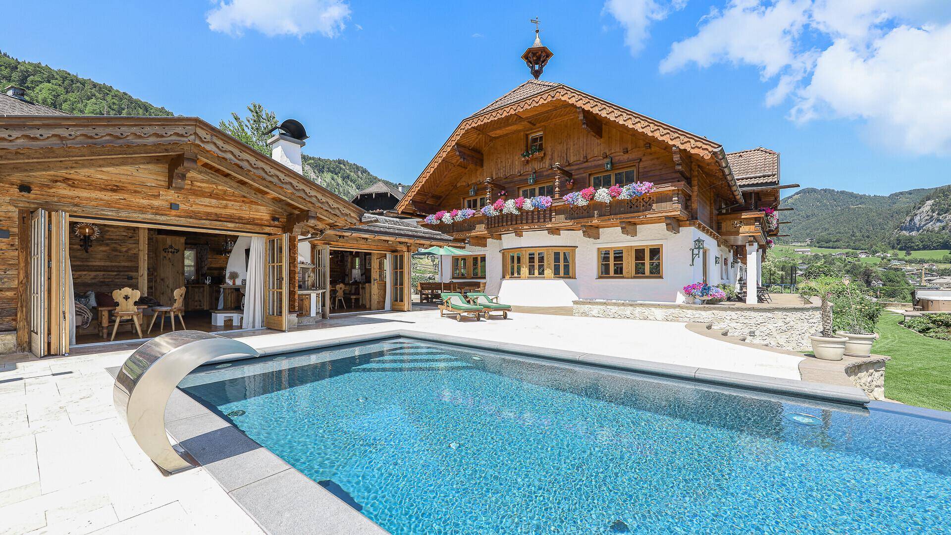 Außenbereich mit Pool, Sonnenterrasse und angrenzendem Holzhaus in alpiner Umgebung.
