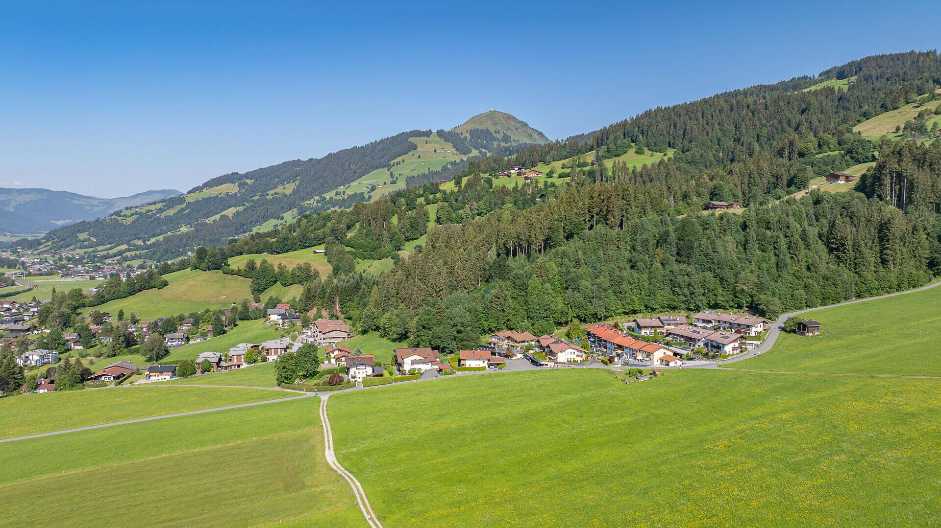 Weitläufige Luftaufnahme eines Tals mit einem Dorf, umgeben von grünen Wiesen und dichten Wäldern.