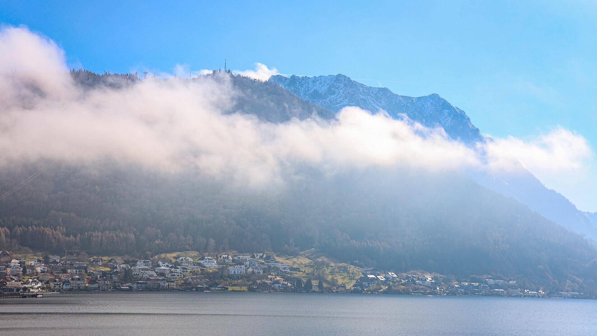 Blick auf eine kleine Stadt am Ufer eines Sees, umgeben von bewaldeten Bergen und Wolken.