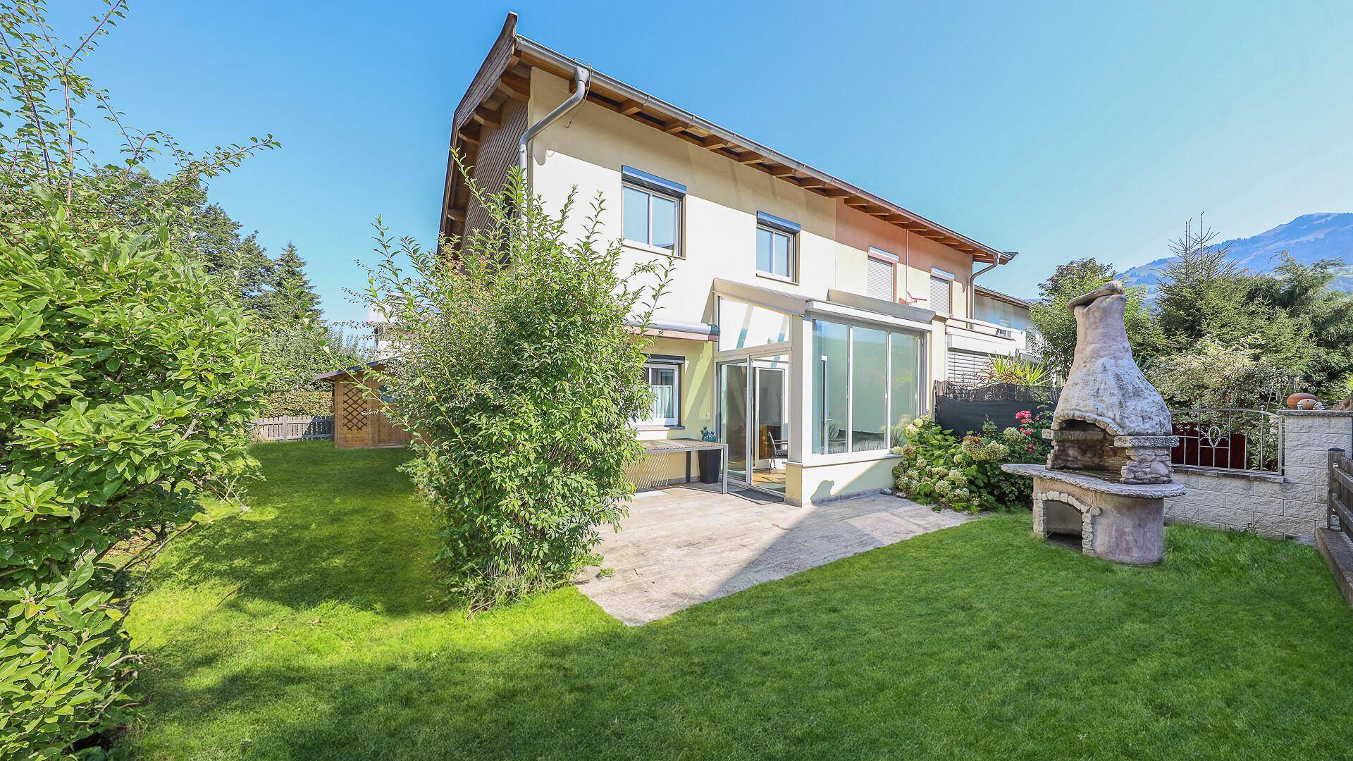 Sonniger Gartenbereich mit Rasen, Terrasse, Wintergarten und traditionellem Grillplatz.