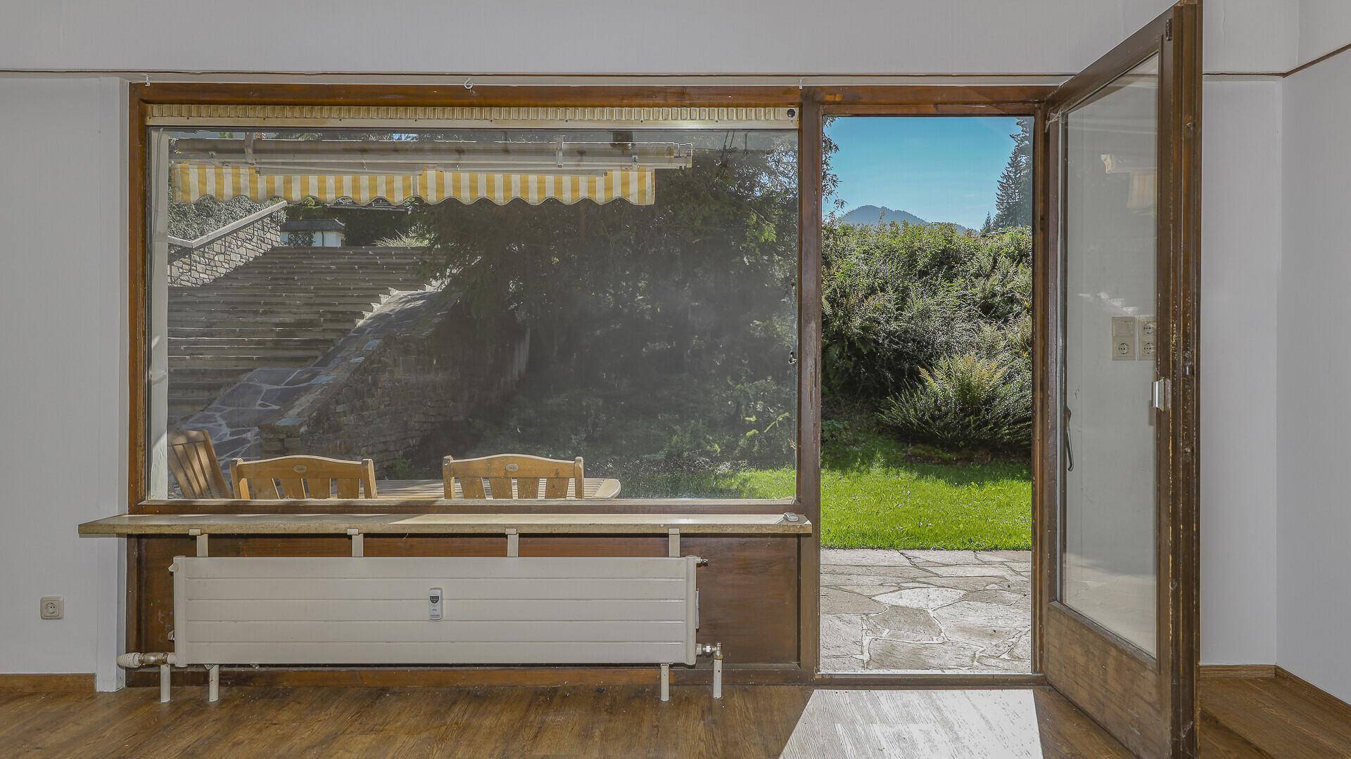 Heller Raum mit Holzboden, großem Fenster und Zugang zu einer Terrasse mit Steintreppe.