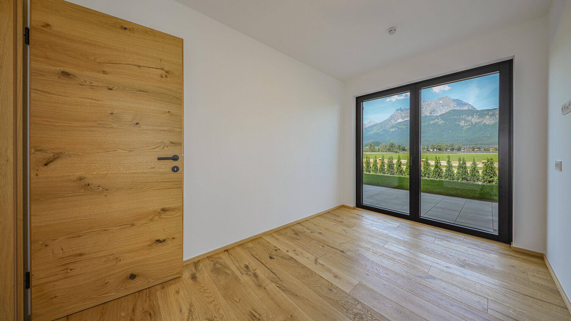 Heller, unmöblierter Raum mit Holztür, Holzboden und Bergblick durch große Fenster.