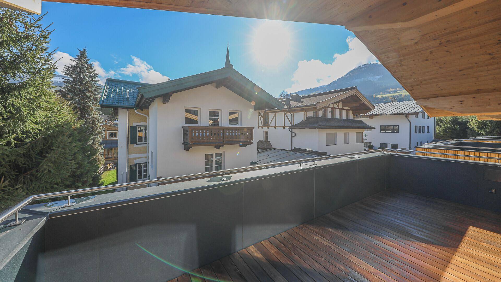 Sonniger Balkon mit Holzboden und Blick auf traditionelle Häuser und die umliegende Berglandschaft.