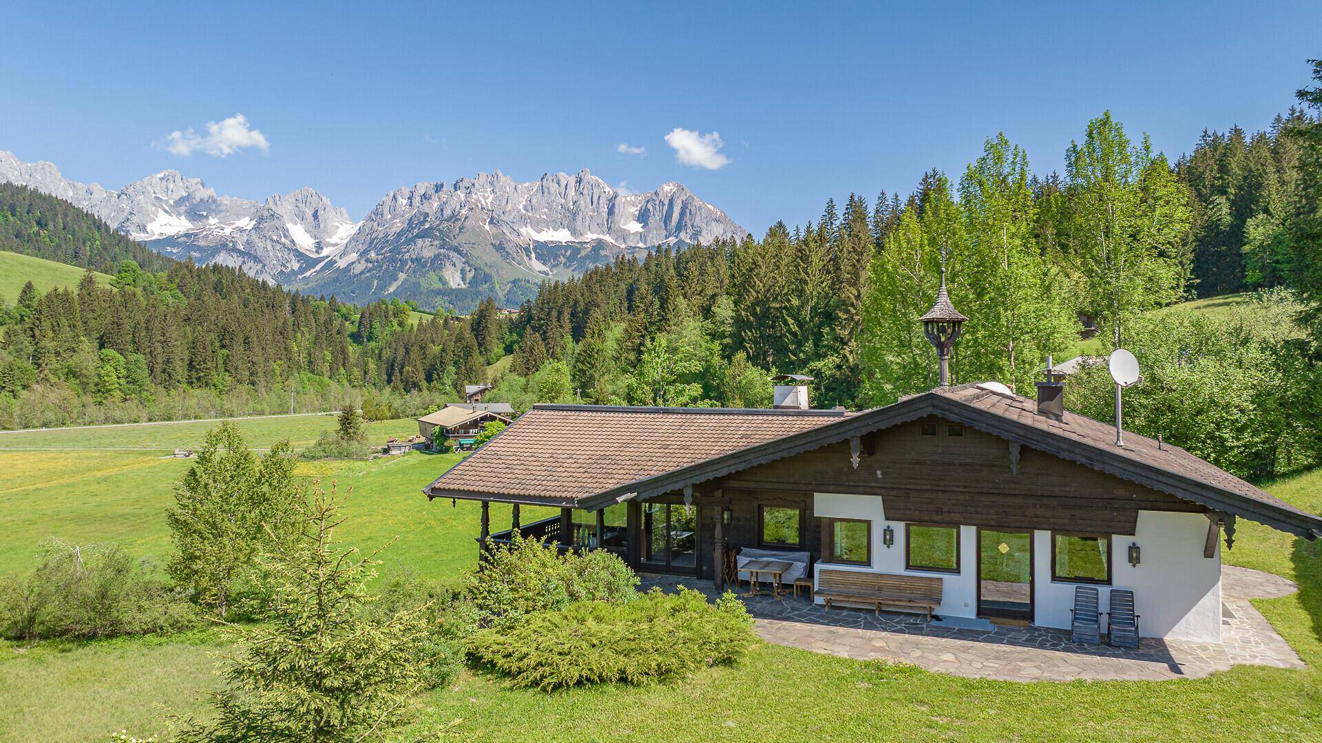 Alpenländisches Haus in sonniger Lage mit Bergblick, umgeben von grünen Wiesen und Wäldern.