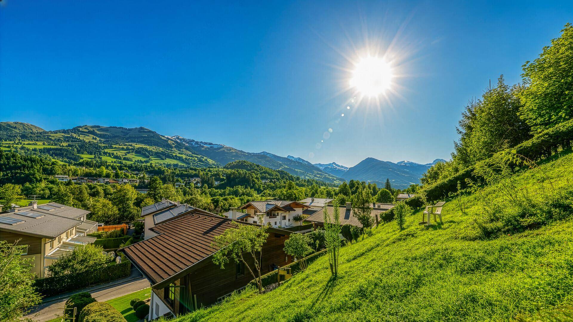 Sonniger Ausblick von einem Hang auf Häuser, grüne Hügel und ferne Berge.