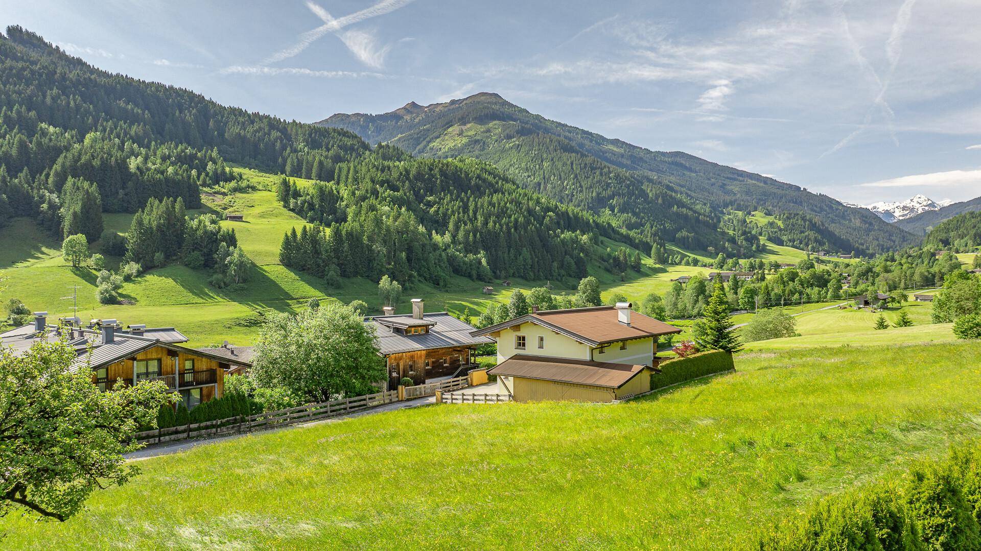 Idyllischer Blick auf ein Haus in einem weiten grünen Tal, umgeben von Wäldern und Bergen.