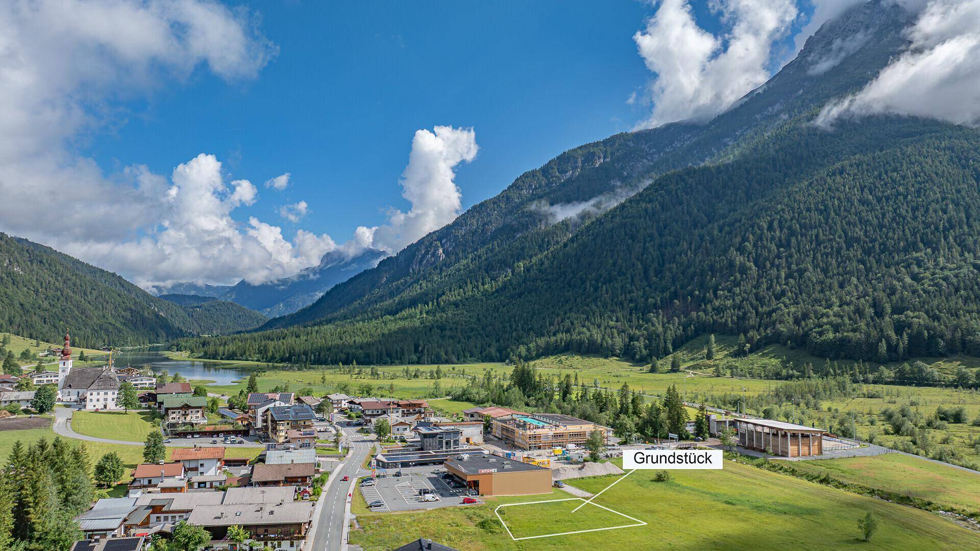 Weitläufige Luftaufnahme eines alpinen Tals mit Dorf, See und markiertem Baugrundstück für Entwicklung.