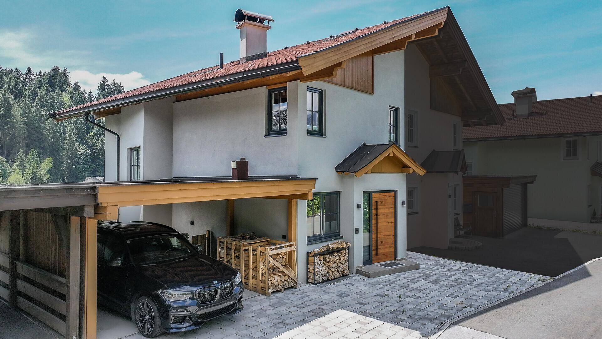 Modernes Einfamilienhaus mit Carport, gepflasterter Auffahrt und Holzstapeln vor alpiner Kulisse.