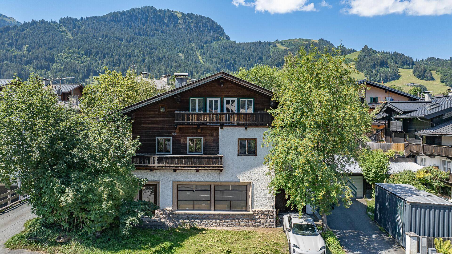 Traditionelles Haus mit Holzbalkonen und weißer Fassade, umgeben von Bäumen und Bergen.