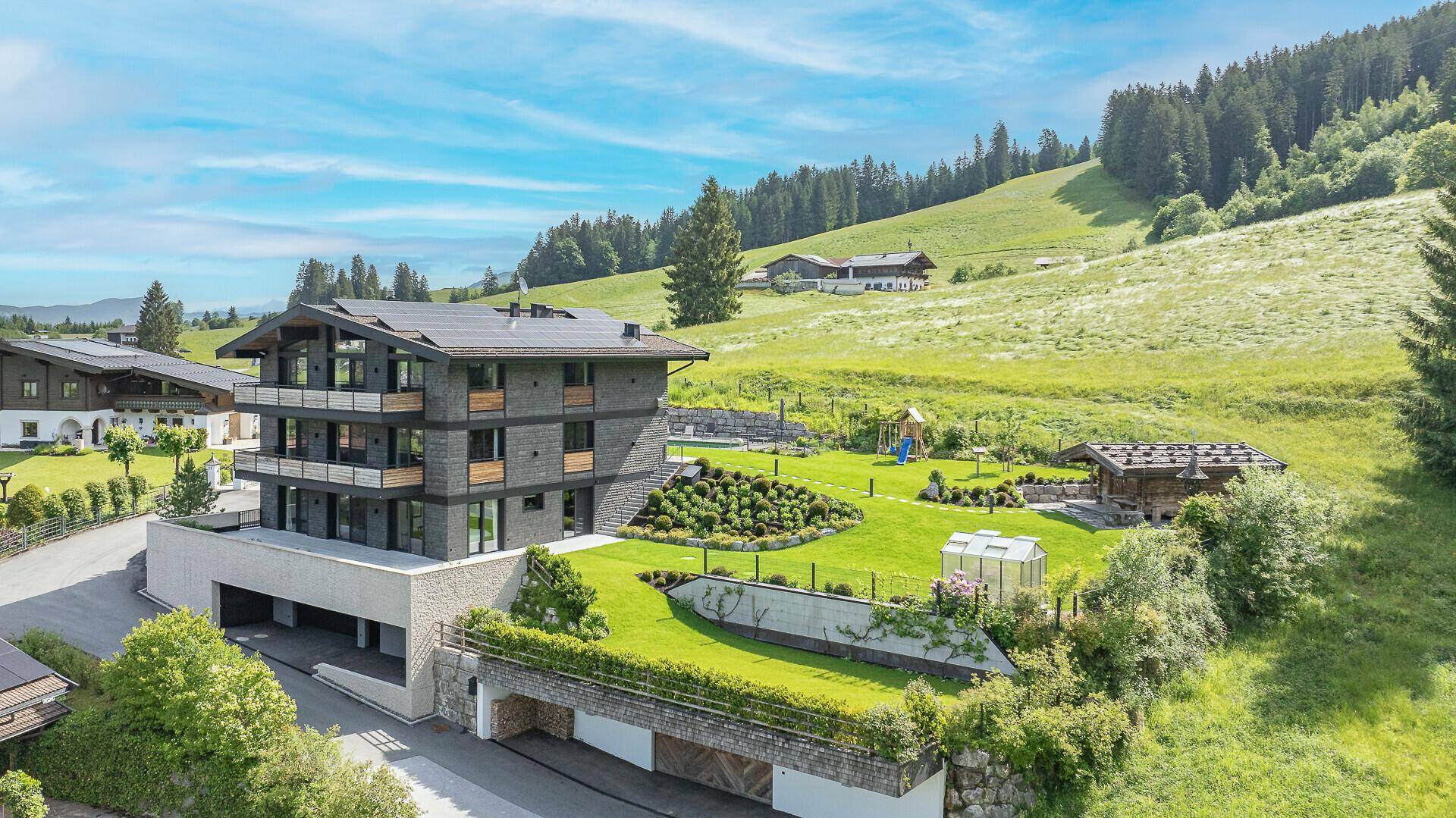 Modernes Mehrfamilienhaus in alpiner Landschaft mit großem Garten und Spielplatz.