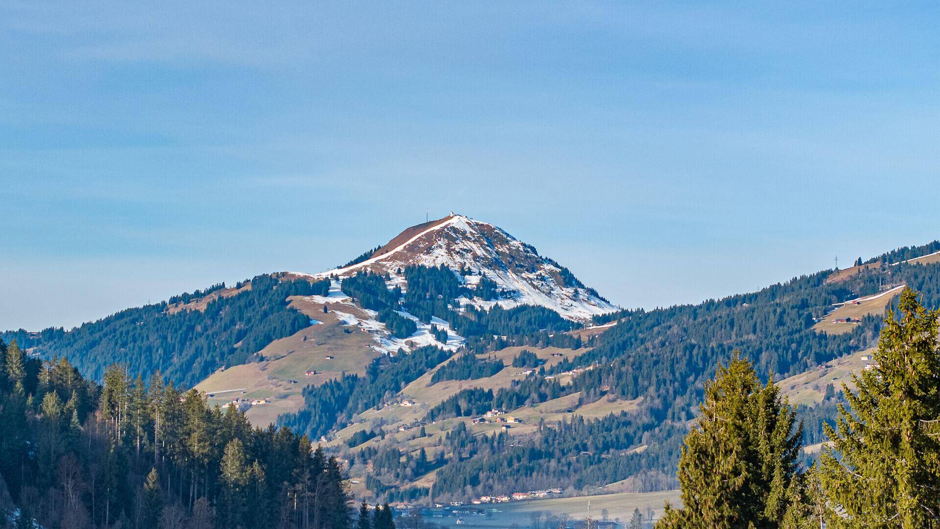 Malerische Berglandschaft mit schneebedeckten Gipfeln und grünen Wäldern unter blauem Himmel.