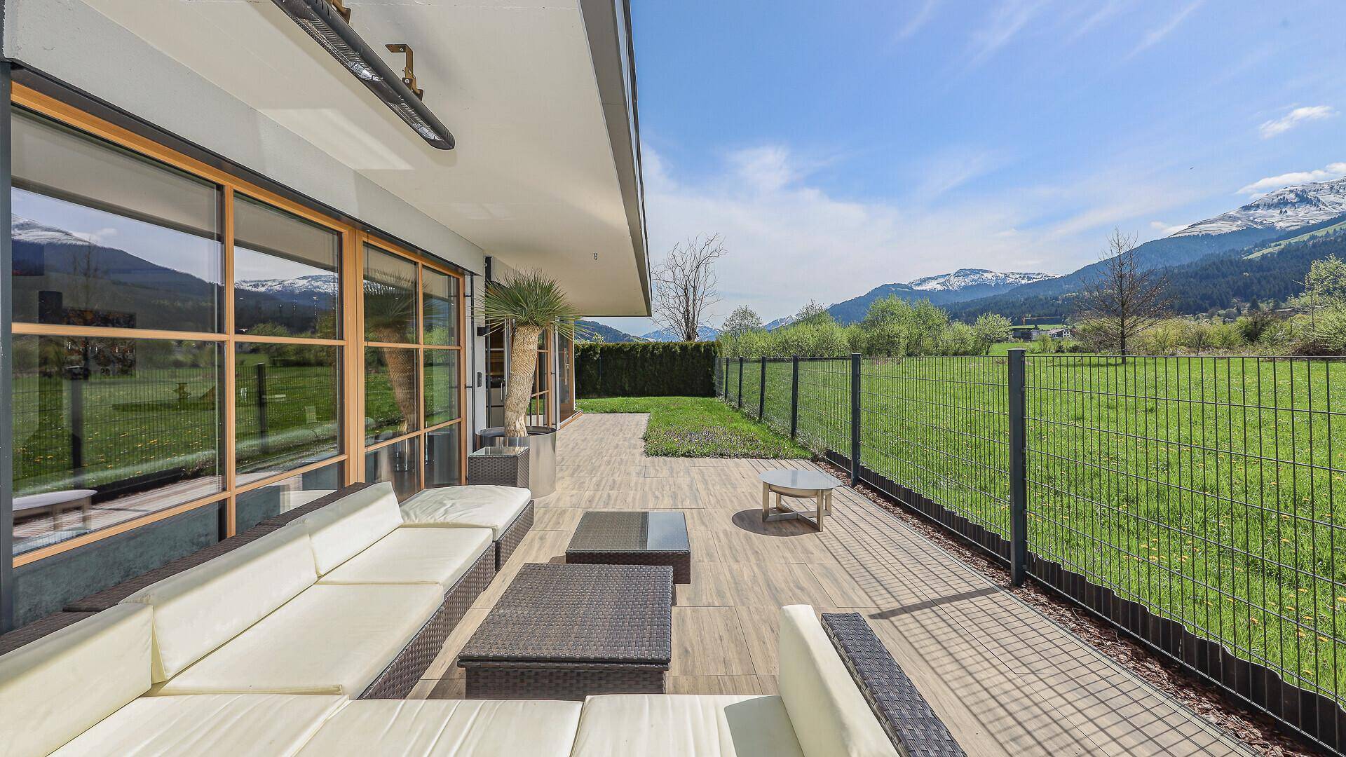 Sonnige Terrasse mit Holzoptik-Fliesen und Zugang zum Garten, umgeben von grüner Natur und Bergen.