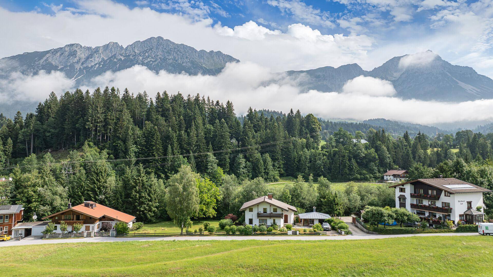 Weitläufige Landschaft mit mehreren Häusern im alpenländischen Stil, umgeben von Wäldern und majestätischen Bergen.