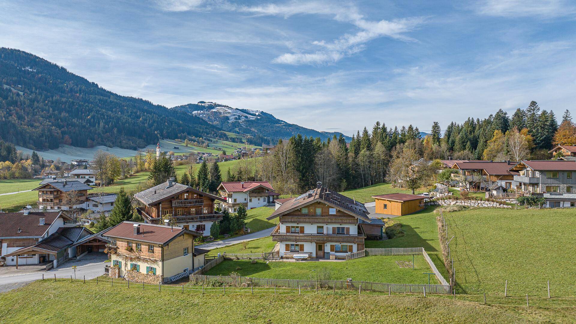 Traditionelles Haus in ländlicher Umgebung mit weiten Wiesen und bewaldeten Bergen im Hintergrund.