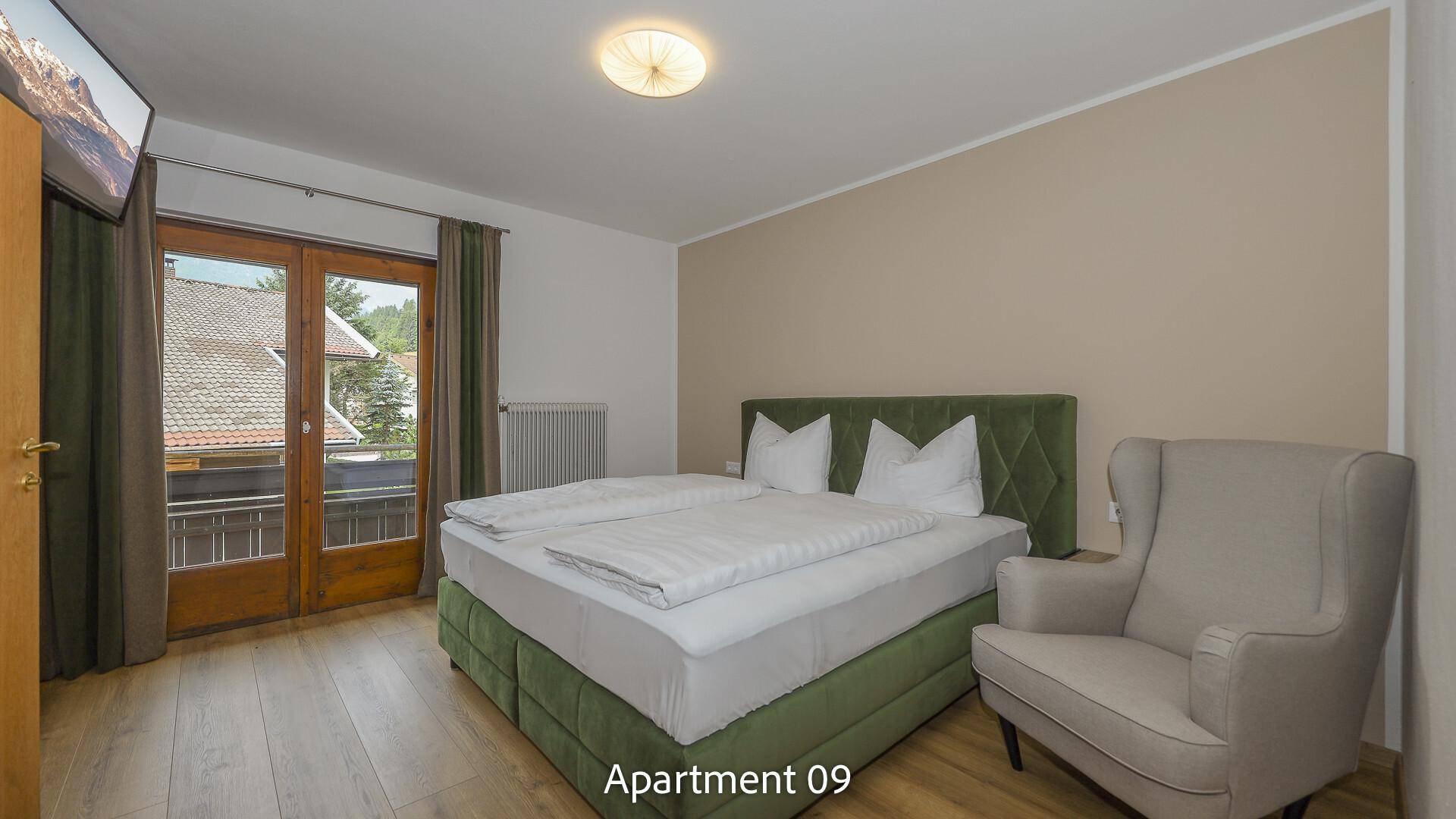 Schlafzimmer von Apartment 09 mit grünem Polsterbett, Sessel und Holzboden, mit Balkonzugang.