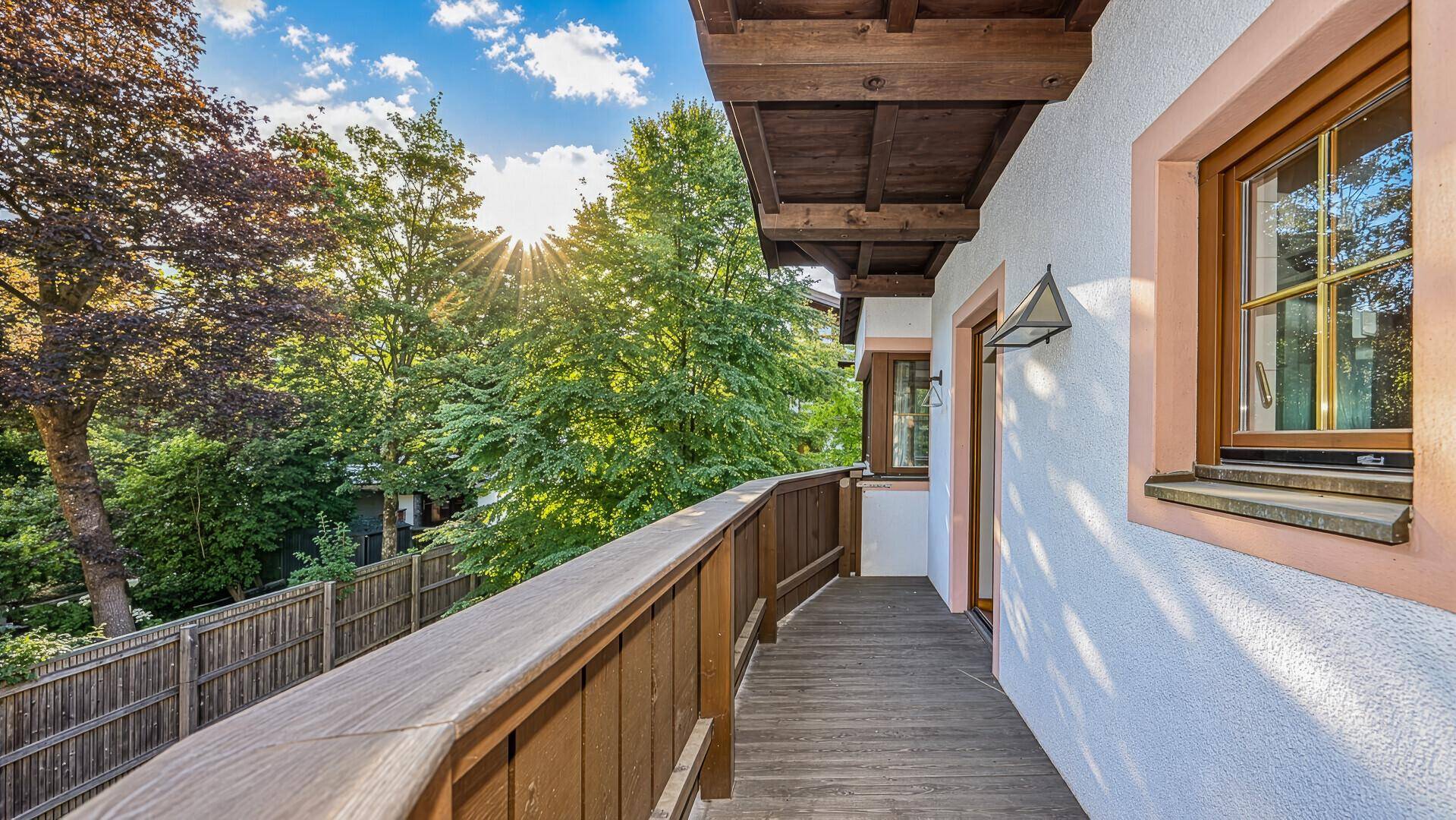 Sonniger Balkon mit Holzboden und Geländer, umgeben von üppigem Grün und Bäumen.