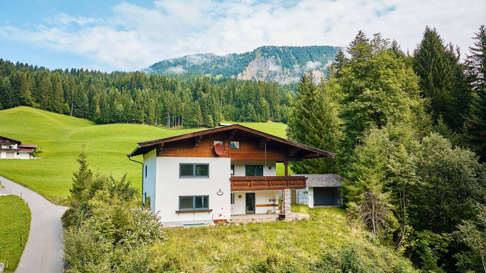 Traditionelles Haus mit Holzbalkon und weißer Fassade, umgeben von grüner Natur und Bergen.