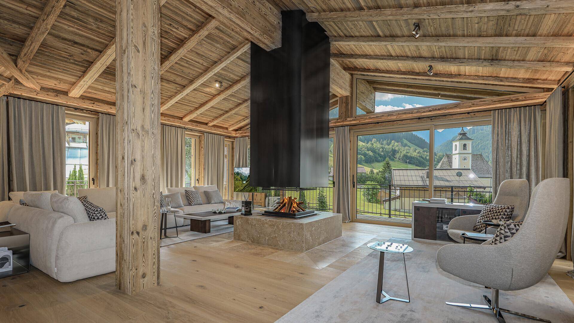 Lichtdurchflutetes Wohnzimmer mit Kamin, Holzbalkendecke und Panoramablick auf die Berge und das Dorf.