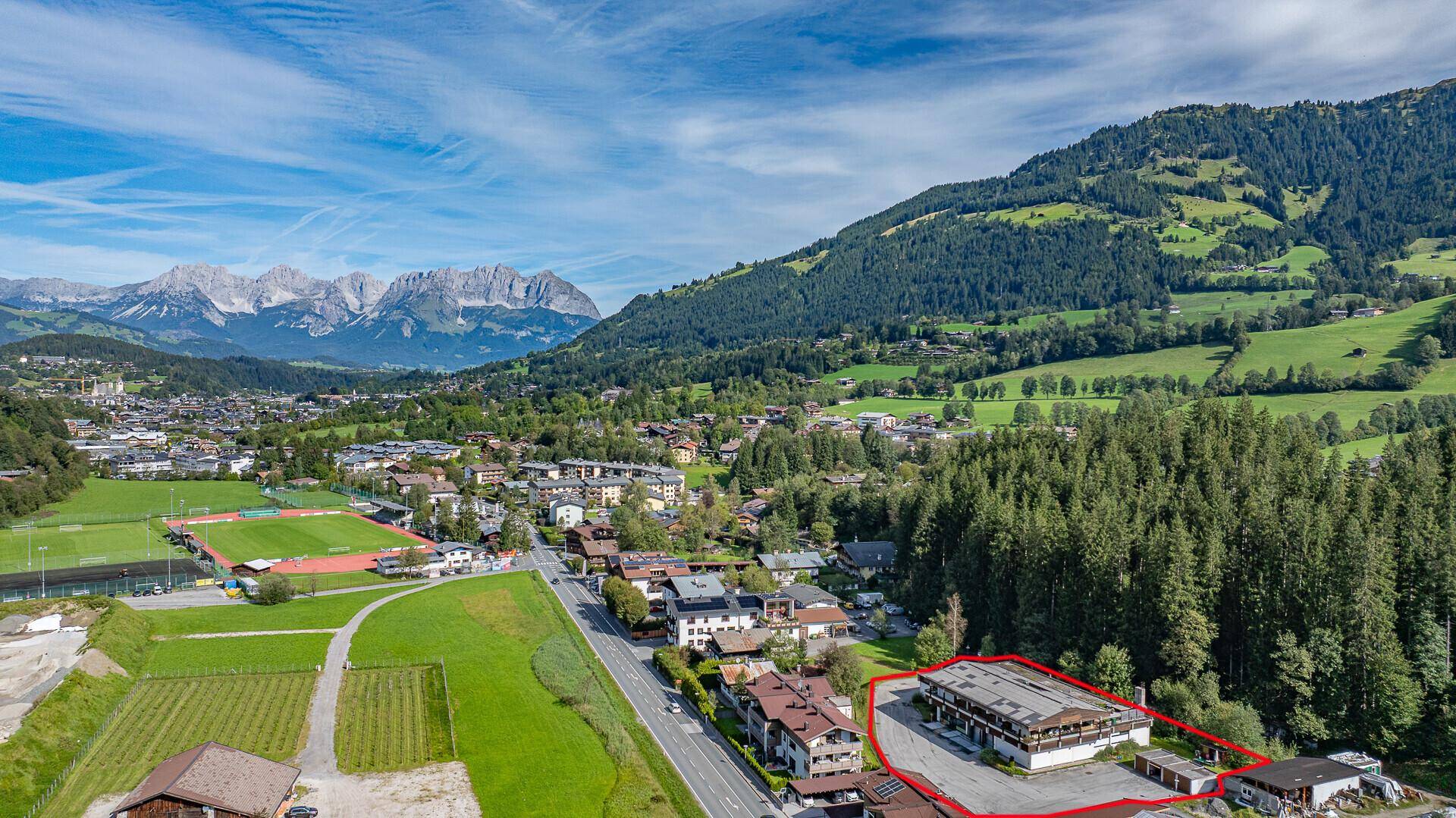Luftaufnahme des umrandeten Grundstücks in einer ländlichen Umgebung mit Blick auf die Berge und ein Sportfeld.