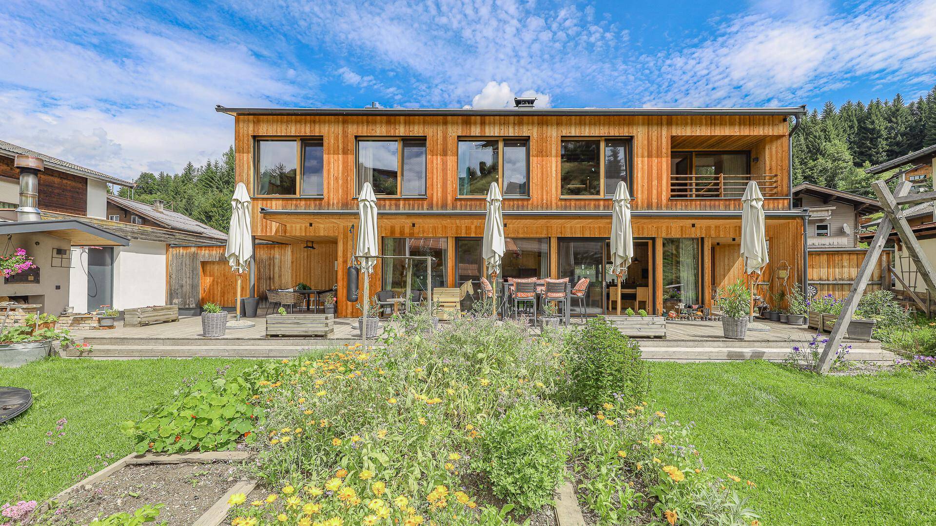Holzhaus mit großer Terrasse und blühendem Gemüsegarten, umgeben von grüner Landschaft.