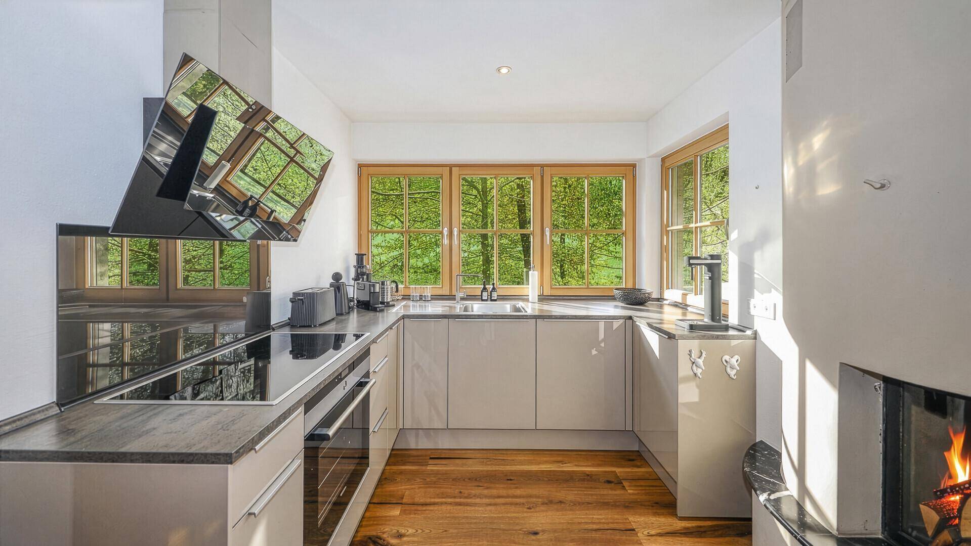 Moderne Küche mit Induktionskochfeld, Backofen und großen Fenstern mit Waldblick.