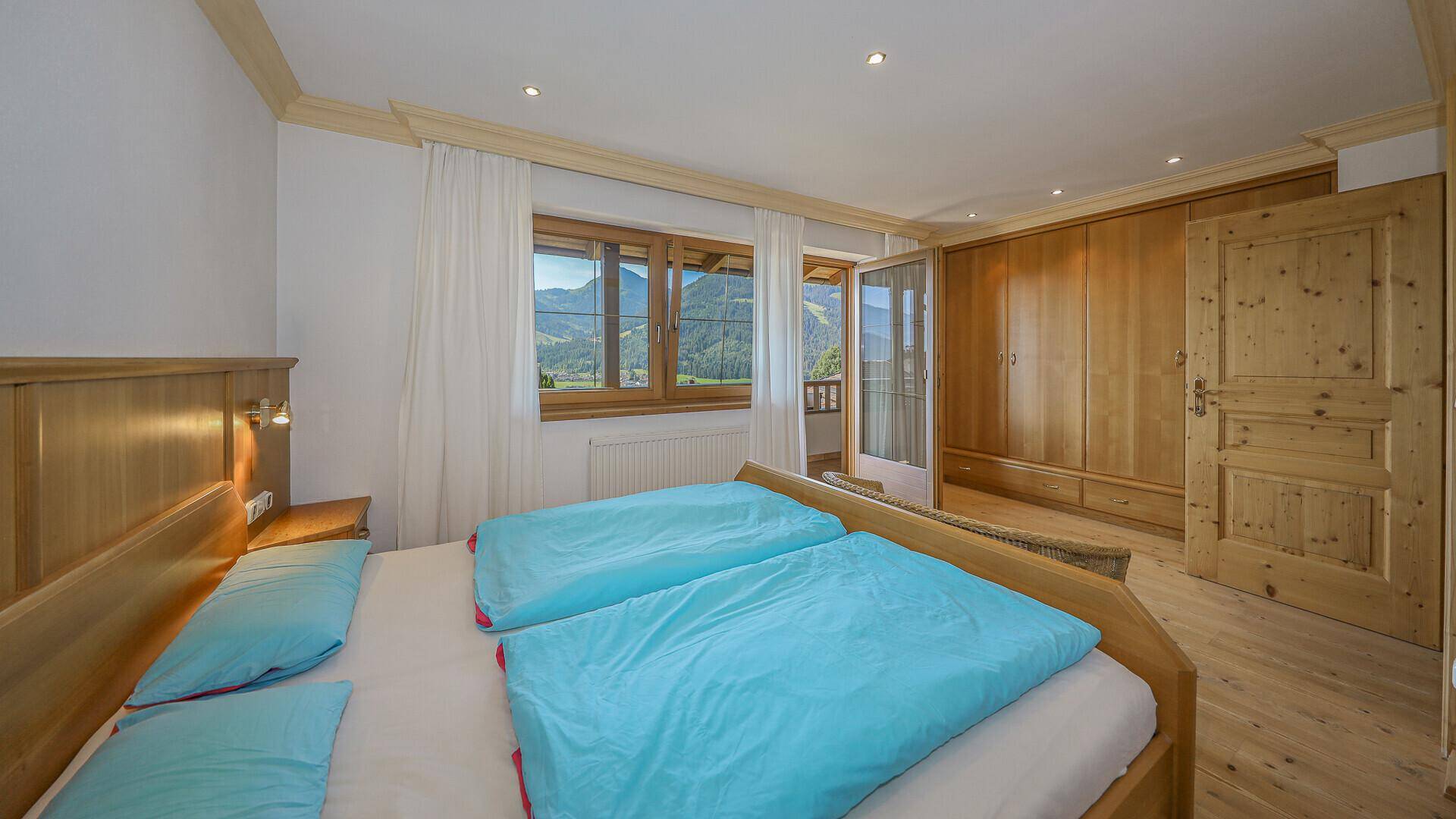 Helles Schlafzimmer mit Doppelbett, Holzmöbeln und Balkonzugang mit Bergblick.