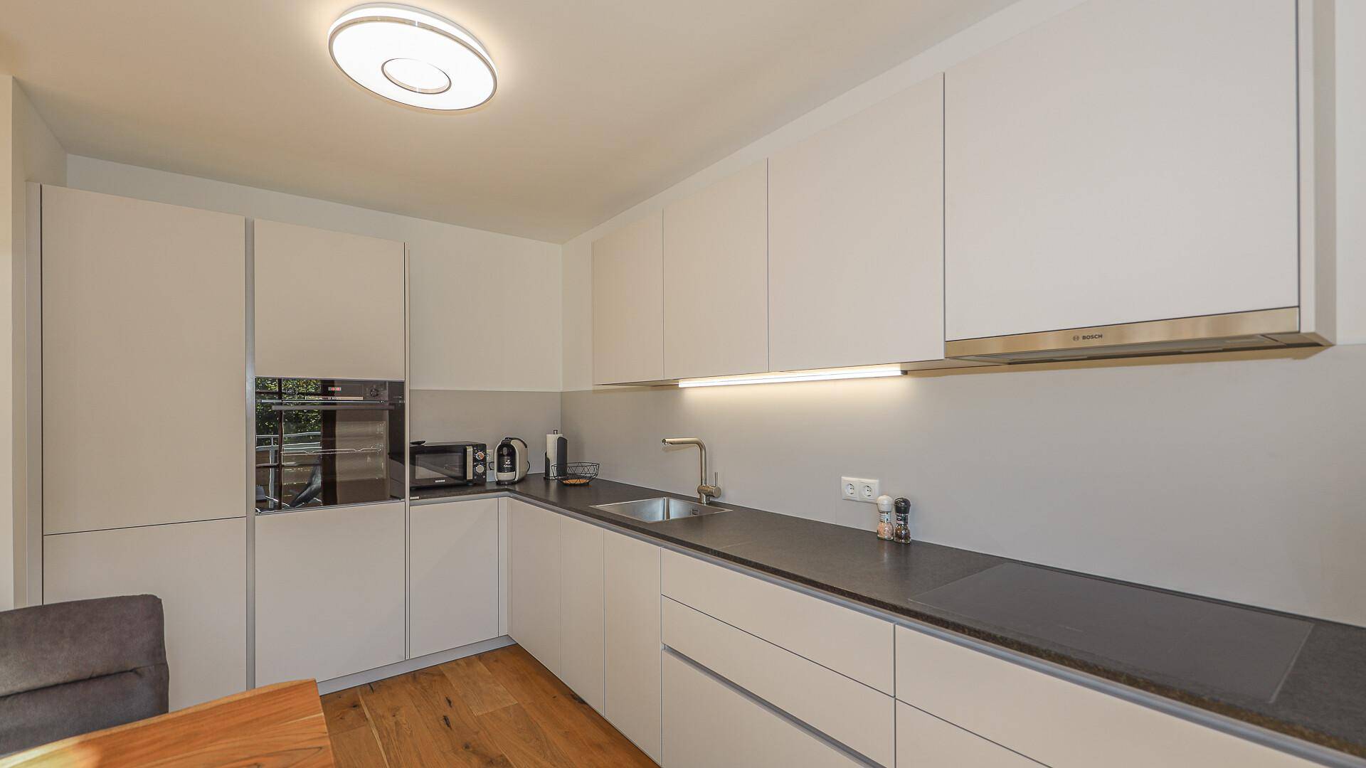 Moderne Einbauküche mit weißen Fronten, dunkler Arbeitsplatte und integrierten Geräten.