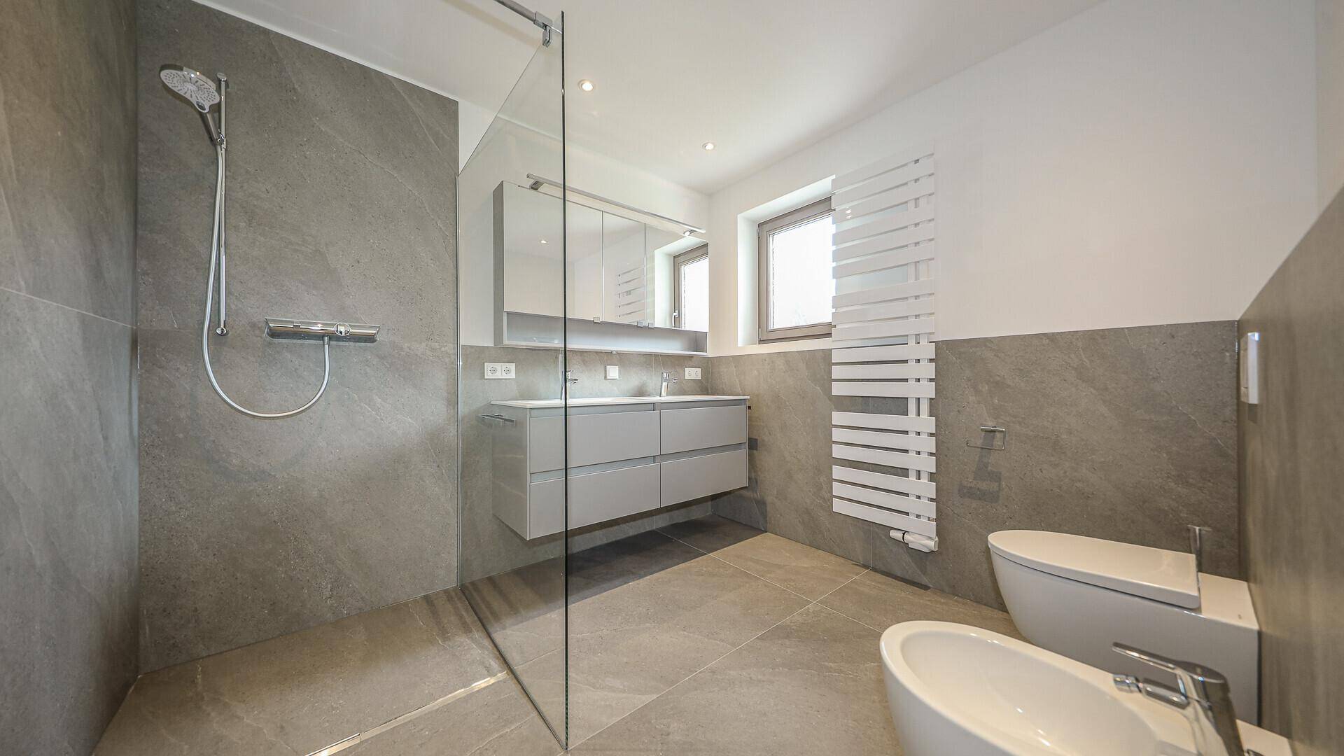 Modernes Badezimmer mit grauen Fliesen, ebenerdiger Dusche, Doppelwaschbecken und Fenster.