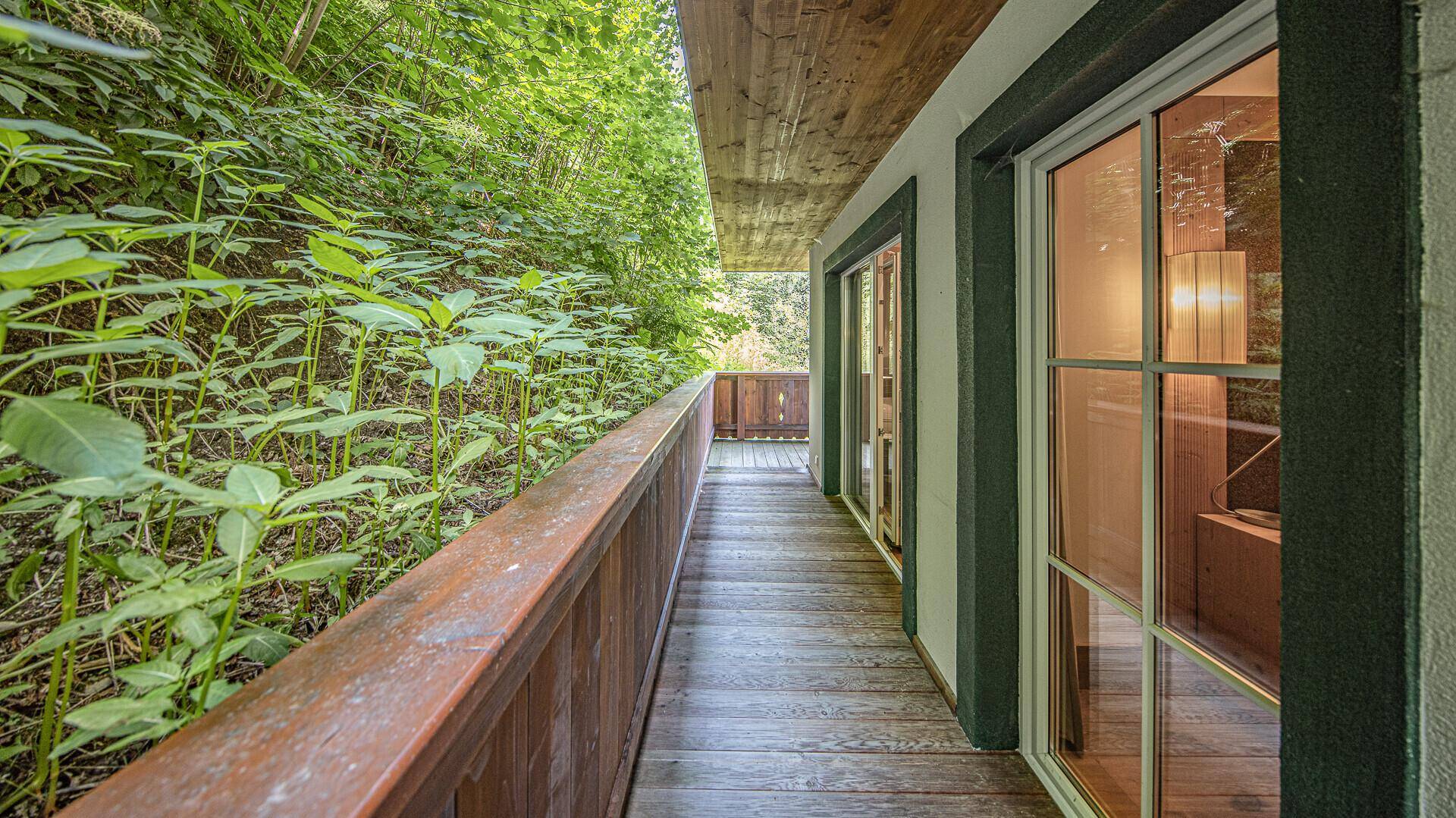 Schmaler Holzbalkon entlang der Fassade mit üppigem Grün und Zugang zu Innenräumen.
