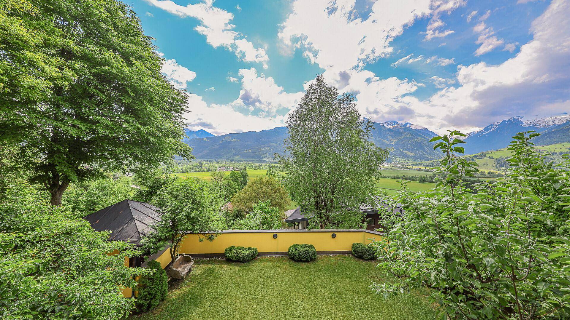 Erhöhter Blick auf den gepflegten Garten mit Laube, gelber Mauer und Bergsicht.