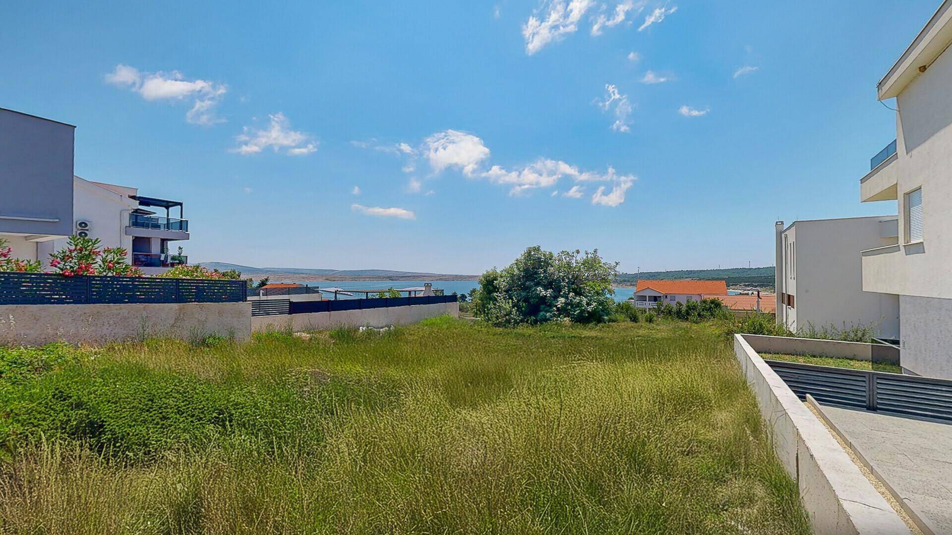 Unbebautes Grundstück mit Gras und Büschen, umgeben von modernen Häusern und Meerblick in der Ferne.