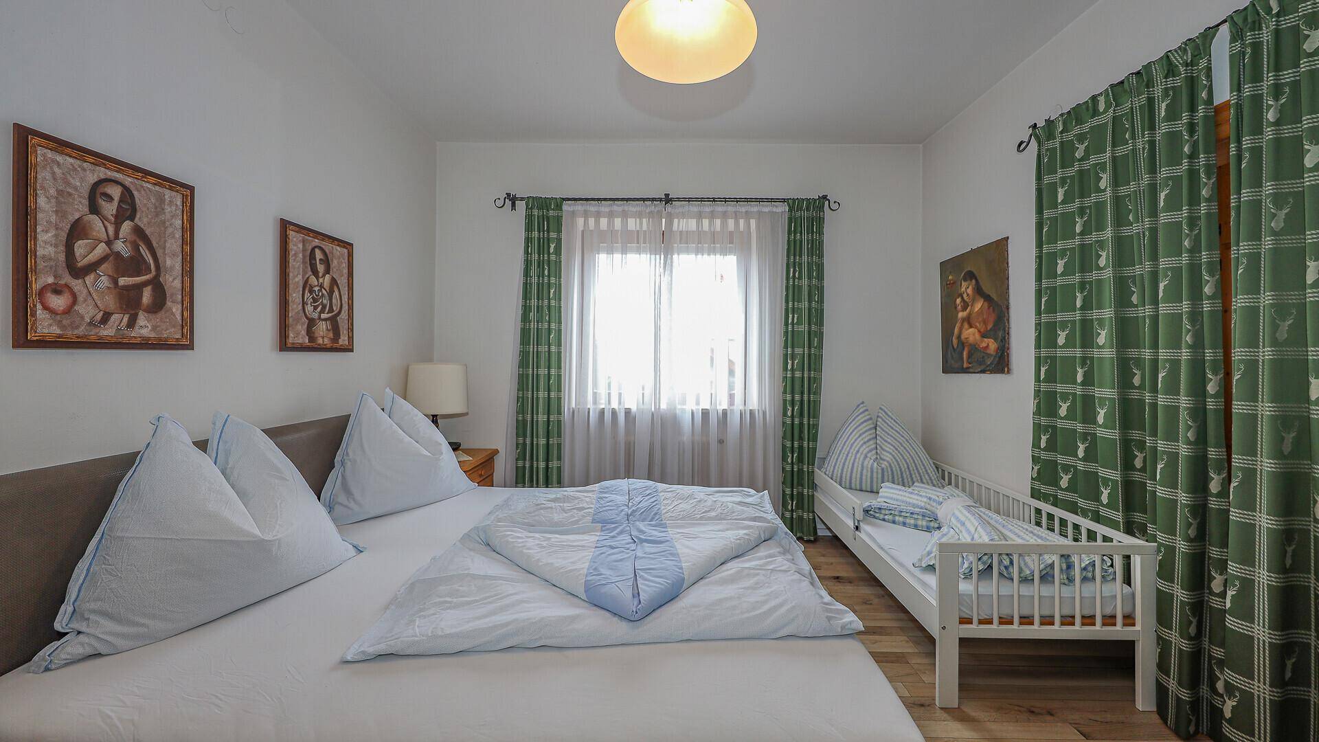 Schlafzimmer mit Doppelbett und Kinderbett, grünen Vorhängen und Blick ins Grüne.