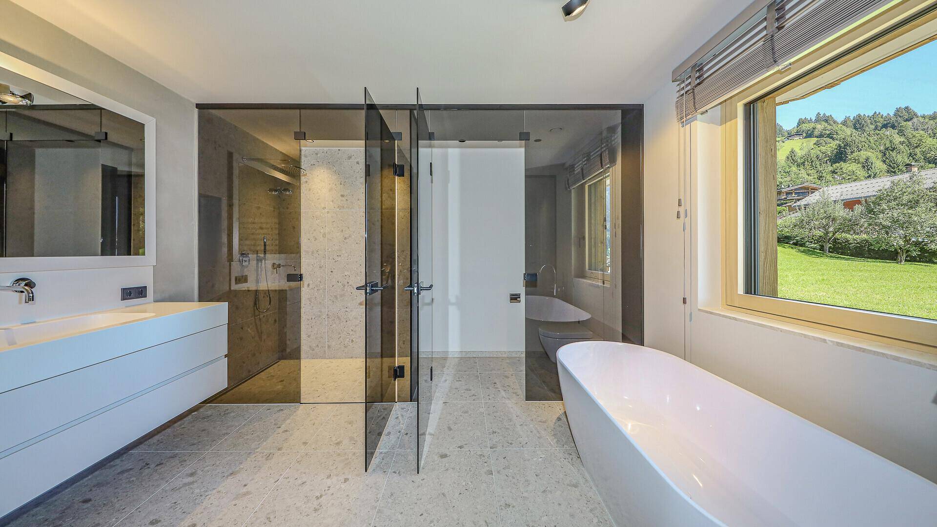 Luxuriöses Badezimmer mit freistehender Badewanne, begehbarer Dusche und Fenster mit Grünblick.