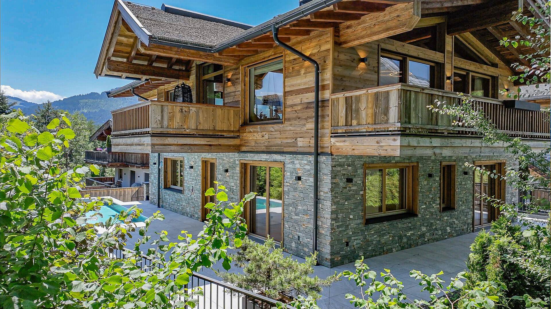 Ansicht des Chalets mit Holz- und Steinfassade, umgeben von üppigem Grün und einem Poolbereich.