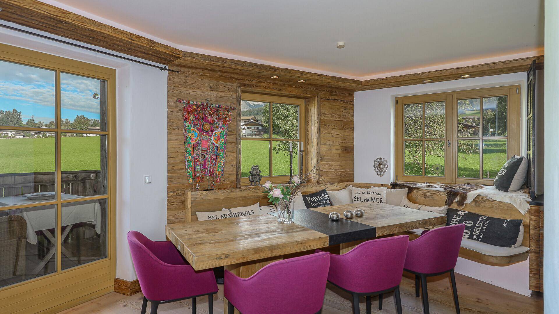 Rustikaler Essbereich mit Holztisch, Sitzbank und Blick auf die grüne Landschaft durch die Fenster.