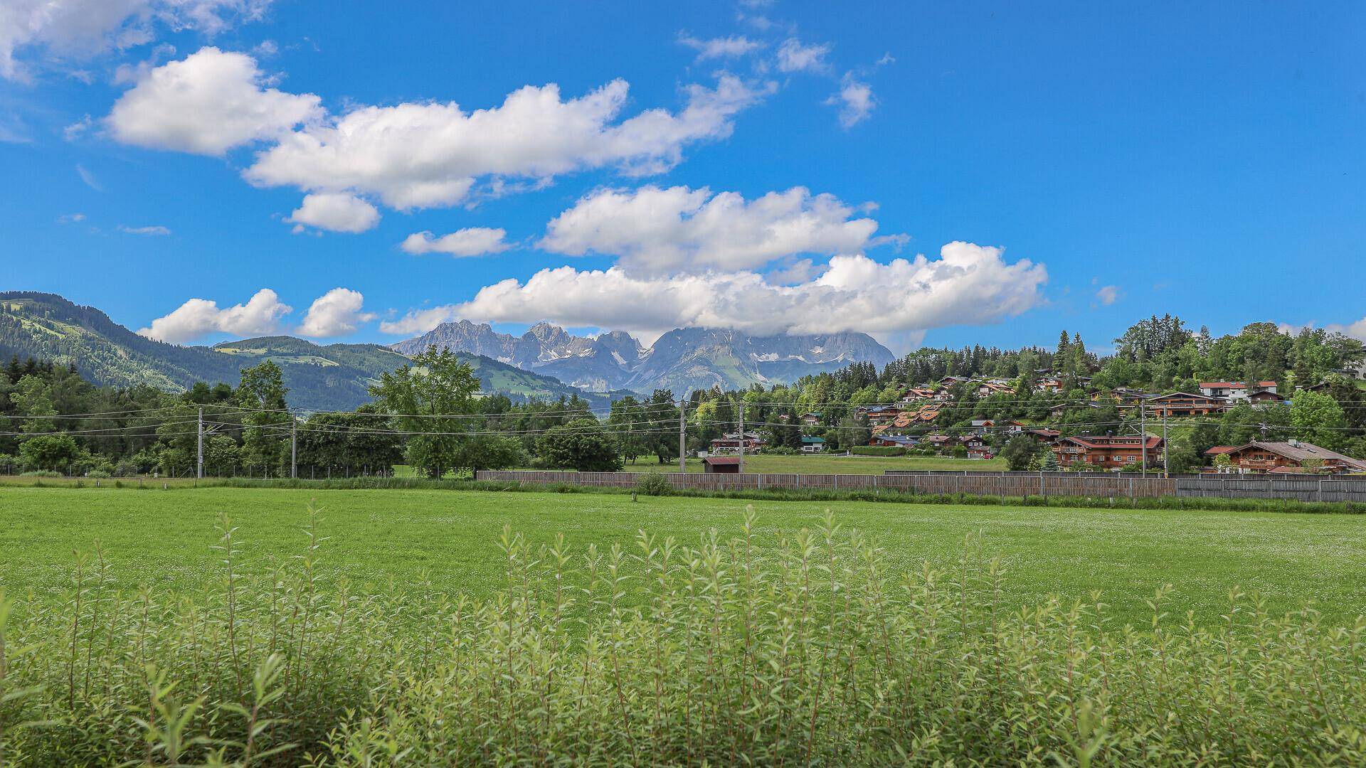 Weitläufige grüne Landschaft mit Wiesen und Wäldern, umrahmt von einer beeindruckenden Bergkette.