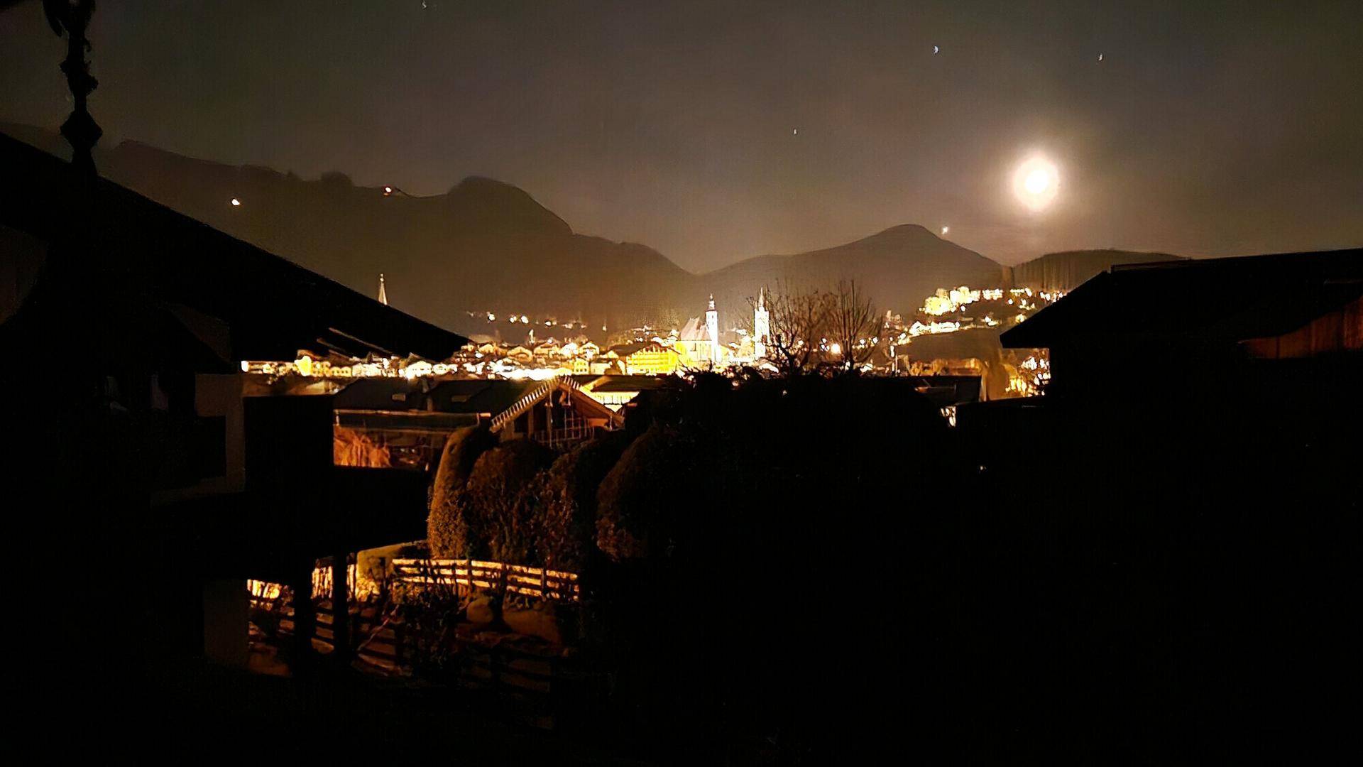 Nachtaufnahme eines beleuchteten Bergdorfes mit Bergen im Hintergrund und hellem Mond am Himmel.
