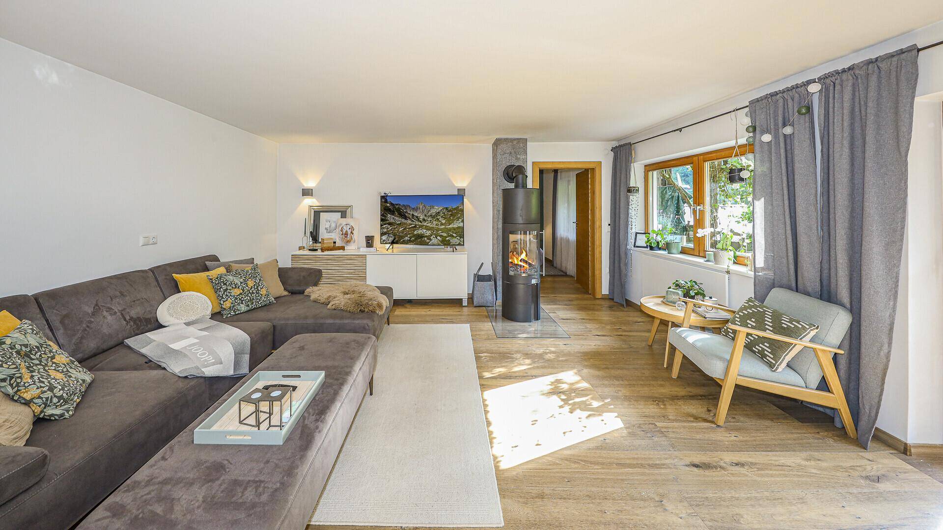 Gemütliches Wohnzimmer mit großem Sofa, Kaminofen und hellen Fenstern, die viel Tageslicht hereinlassen.