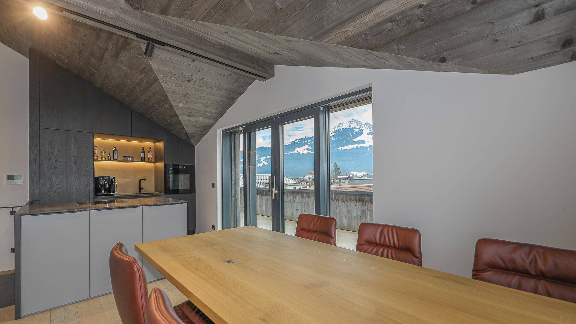 Essbereich mit Holztisch und moderner Küche, umgeben von großen Fenstern mit Bergblick.