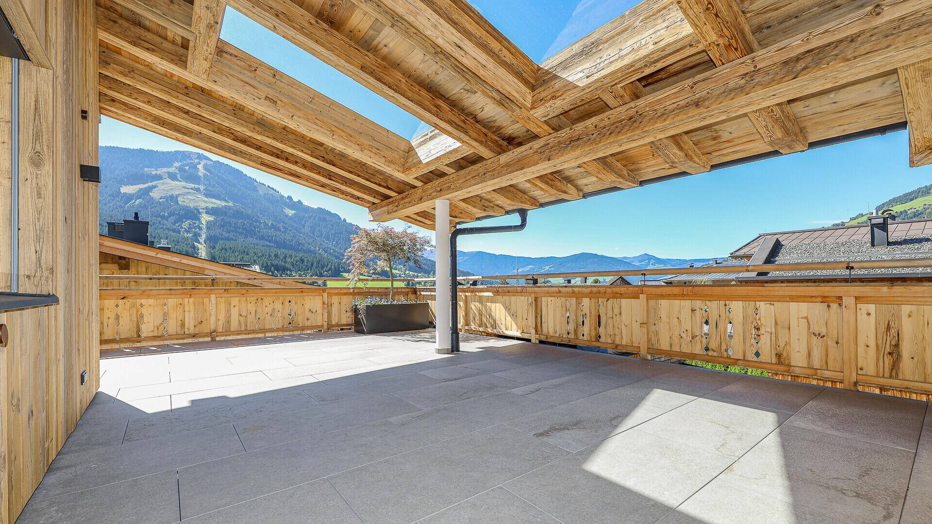 Geräumige Dachterrasse mit Steinplattenboden und Holzbalkenüberdachung, umgeben von Bergen.