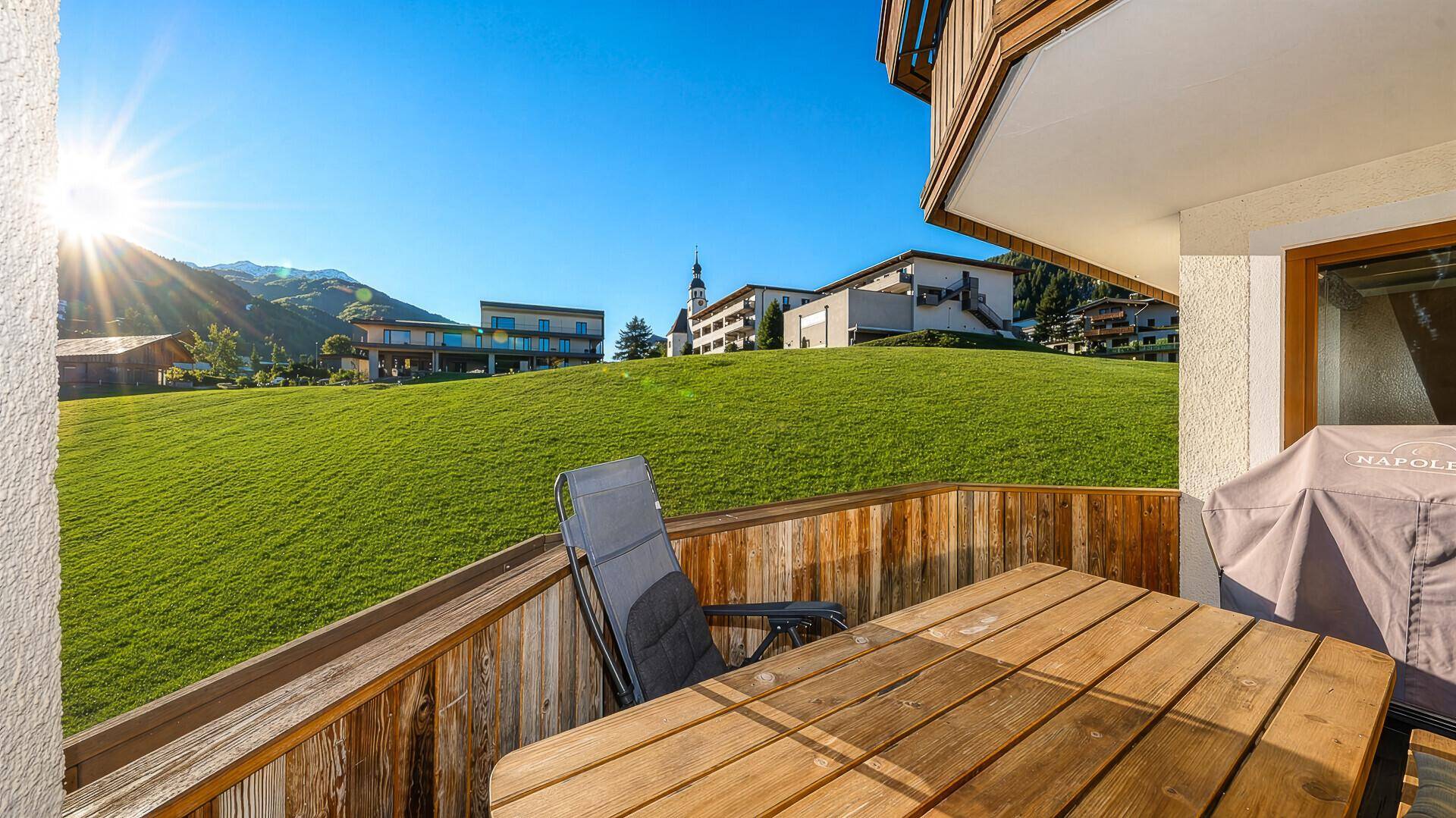 Balkon mit Holztisch und Grill, bietet einen sonnigen Ausblick auf die umliegende Bergwelt.