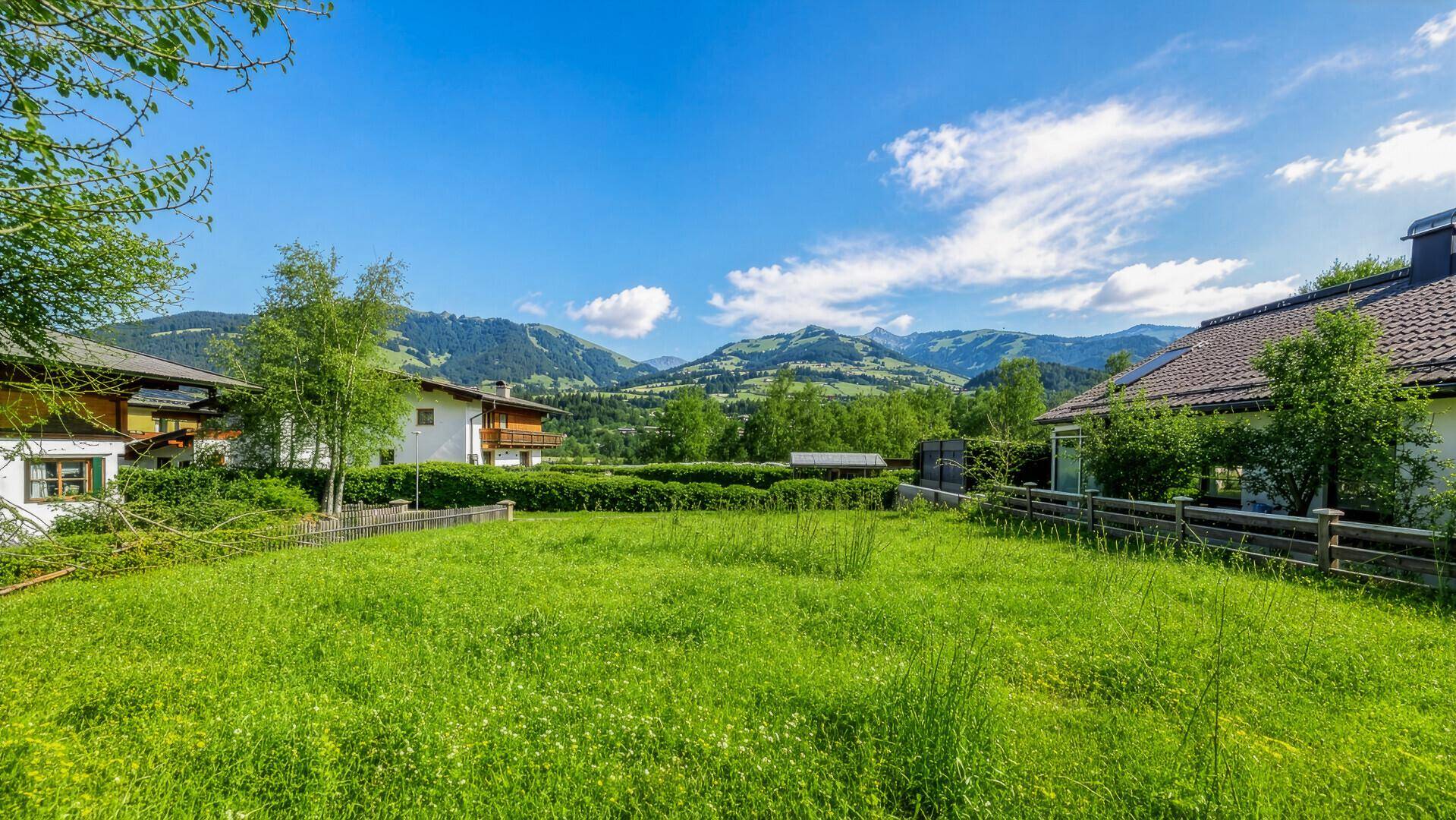 Weitläufige grüne Wiese mit Blick auf traditionelle Häuser und die umliegende Berglandschaft.