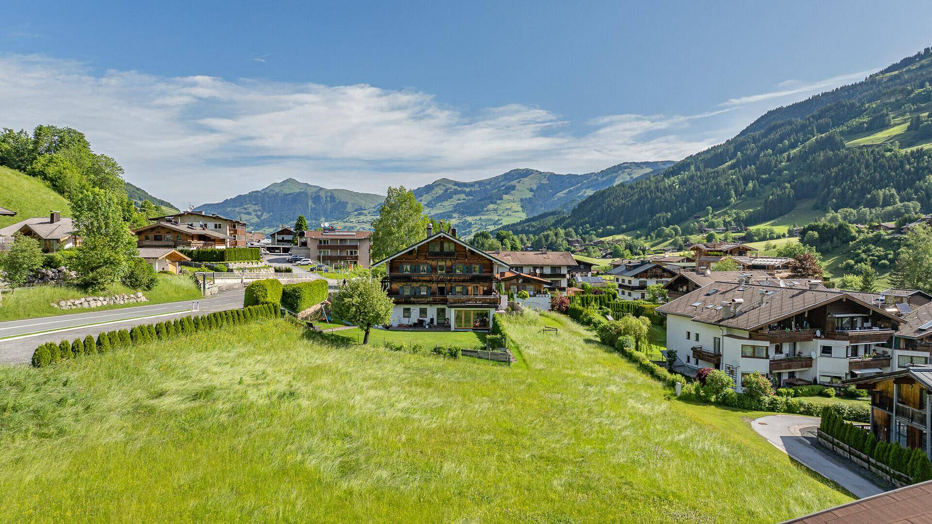 Luftaufnahme eines Dorfes in den Bergen mit grünen Wiesen und umliegenden Häusern unter blauem Himmel.